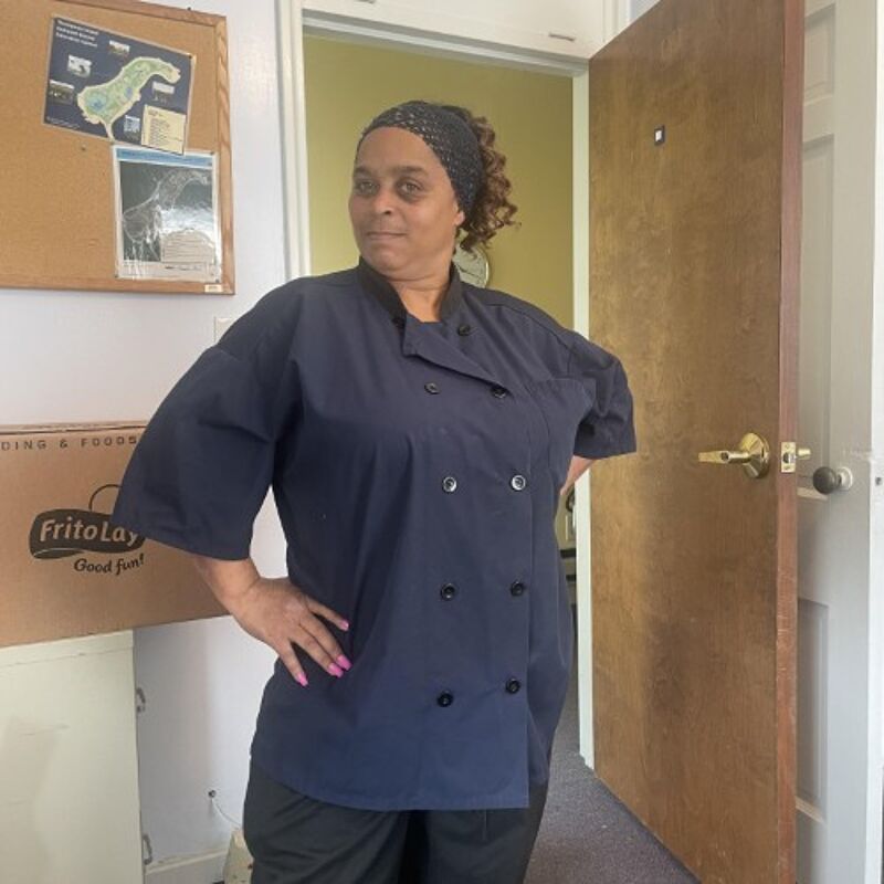 The image shows a woman in a dark blue chef's coat, standing with her hands on her hips. She has a patterned headscarf and is looking at the camera. She is standing in front of a door and a bulletin board with some papers on it. There is a box with the Frito-Lay logo on it.